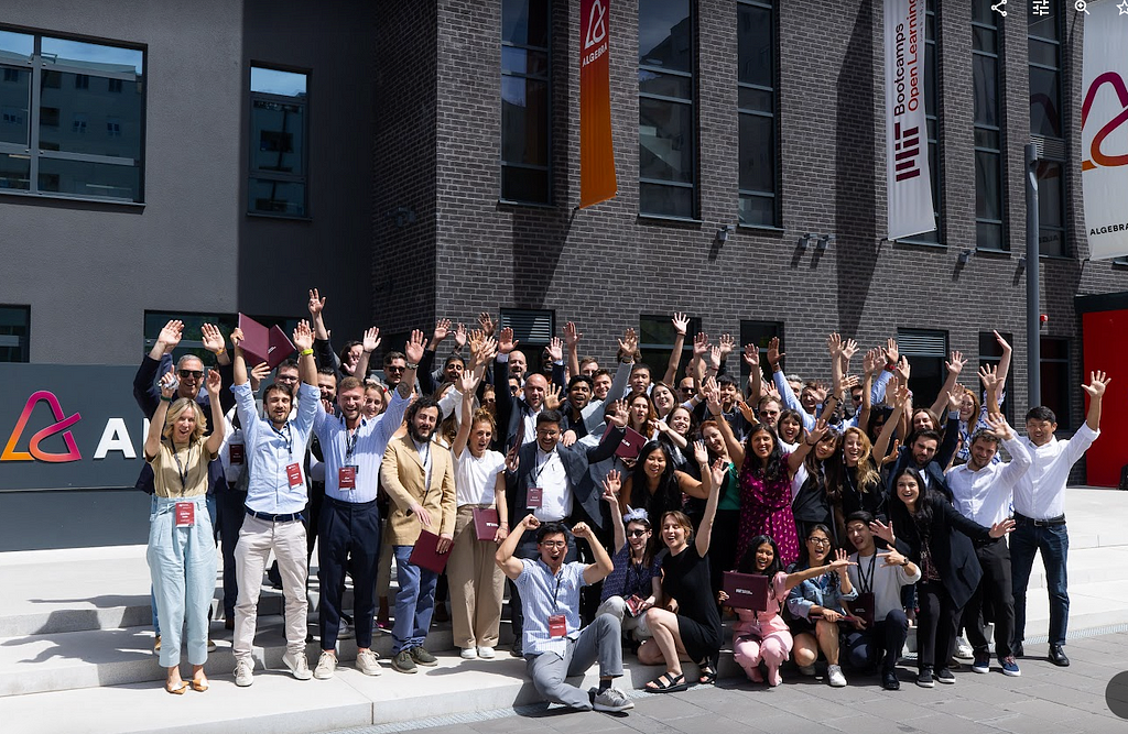 Group photo of approximately 50 adults of different genders and nationalities posing outside of a university building with their hands in the air and cheering.