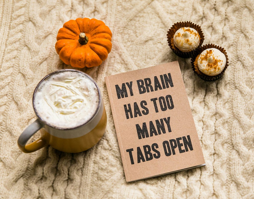 A flat lay of a latte, two cupcakes, pumpkin, and book with a cover that reads “My brain has too many tabs open”.