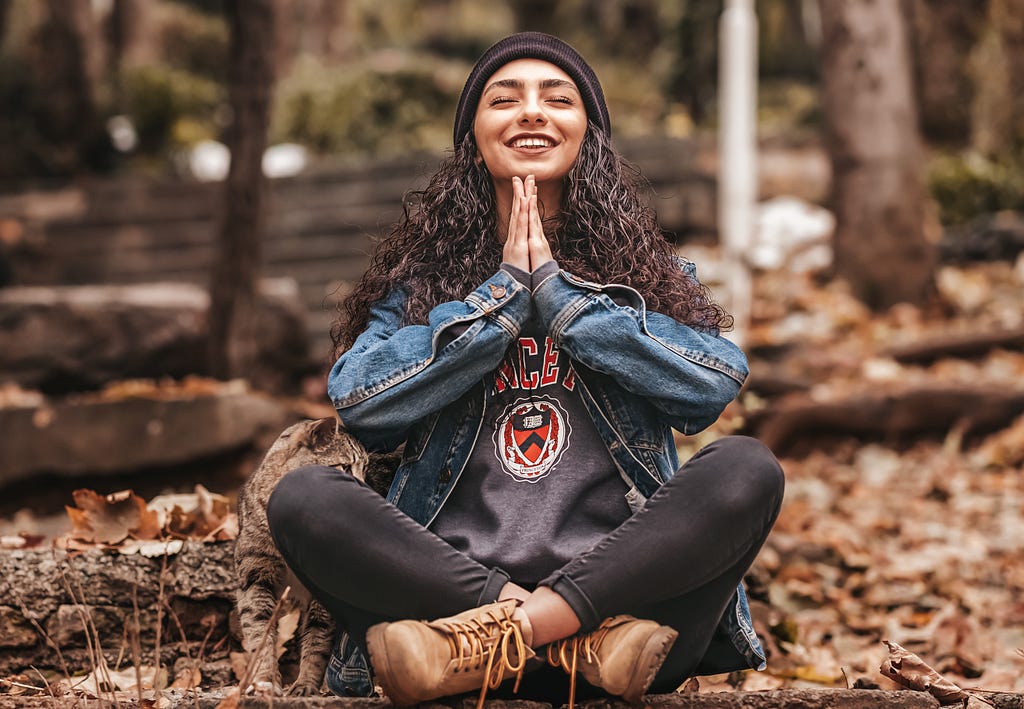 Smiling woman sitting in forest in jean jacket and hiking boots, hands in prayer posture under chin.