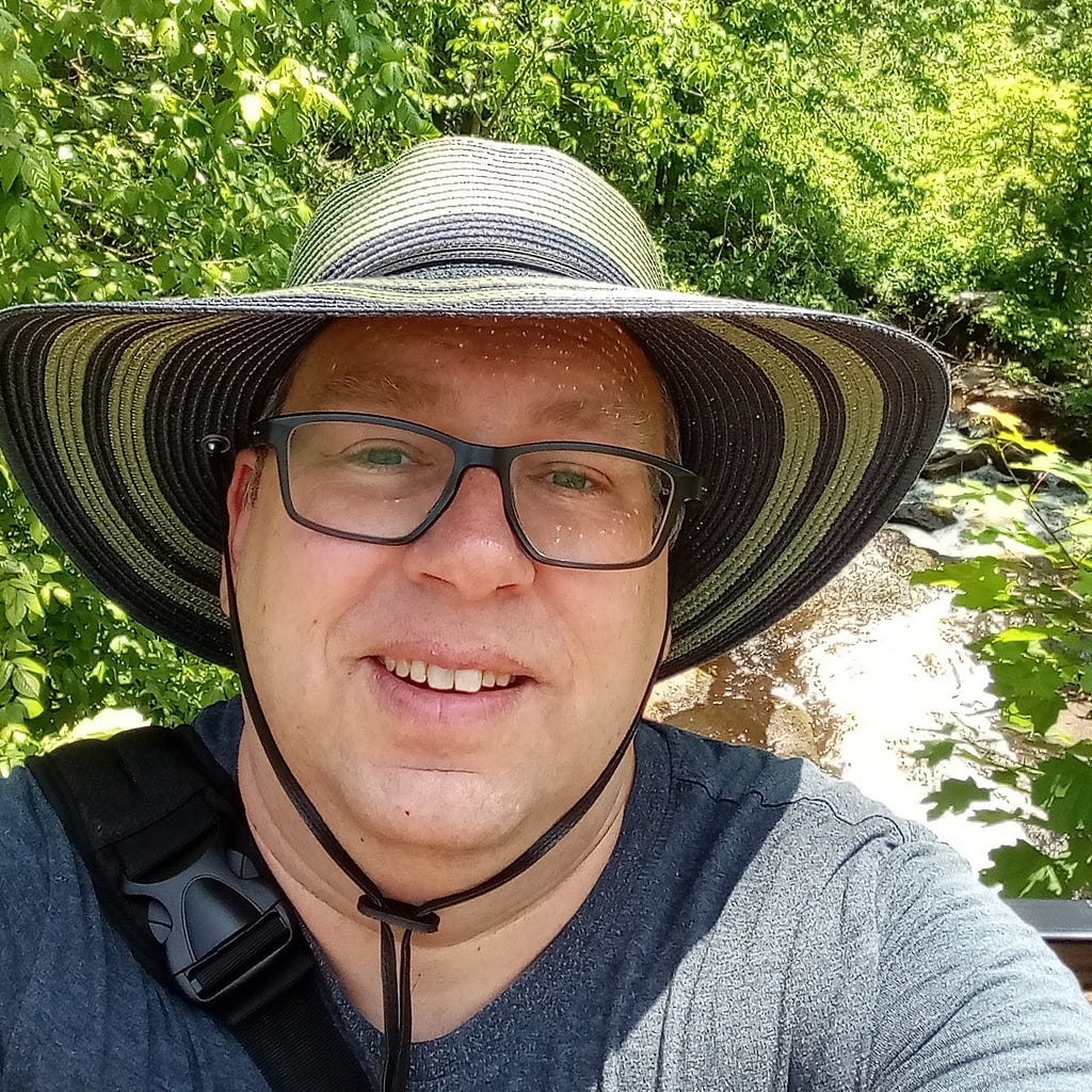 A headshot selfie of the author on a hiking trail wearing a wide-brim hat that could be considered a woman’s hat.