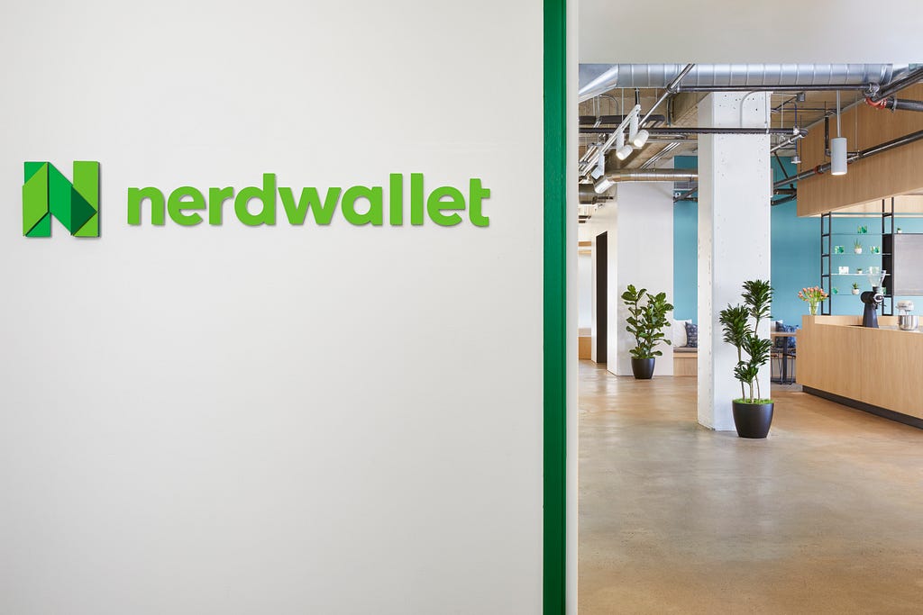 The NerdWallet office entryway. A white wall with the green NerdWallet logo is in focus, and behind it there’s an open concept office space with white pillars, brown floors, and some plants.
