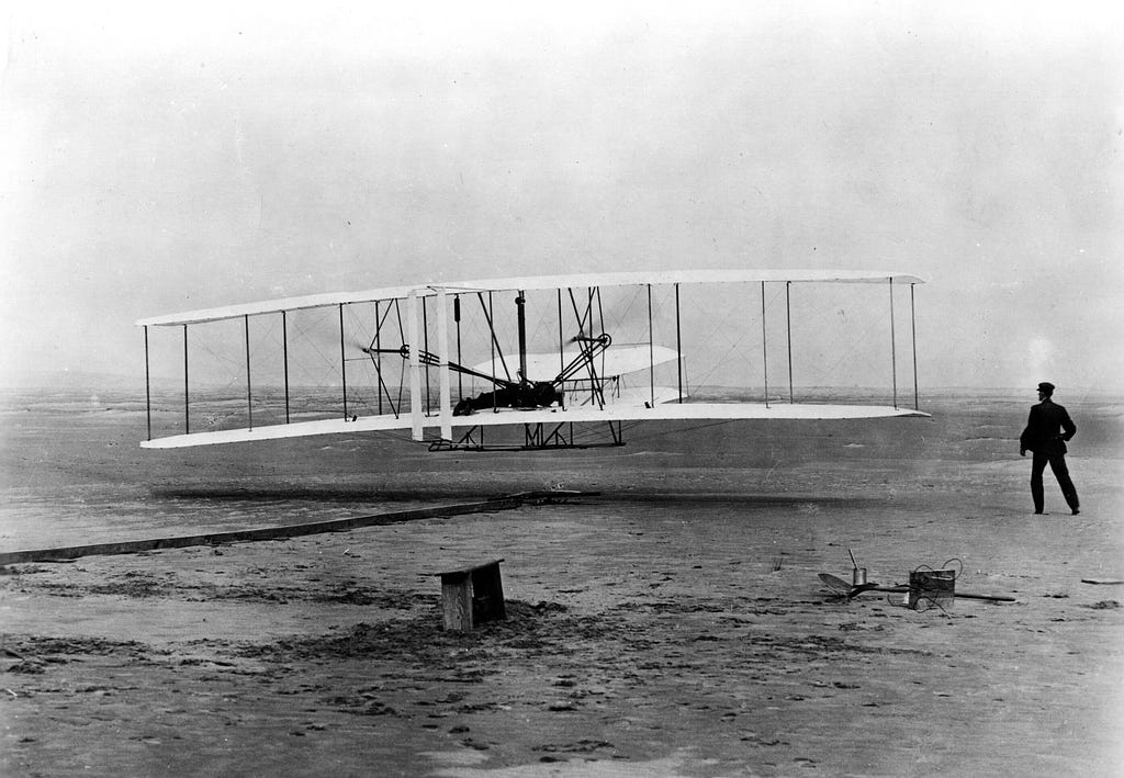 “Wright Brothers’ 1904 Aeroplane (“Kitty Hawk”) in first flight, December 17, 1903 at Kitty Hawk, N.C. Orville Wright at controls. Wilbur Wright standing at right.