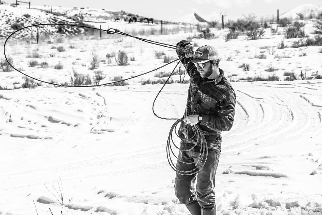 Black and white photo of man twirling a lasso in snow-covered Wyoming.
