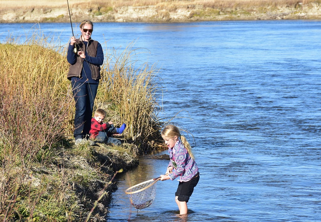 woman and two kids fishing. Adult has rod, one child sits next to her , other in stream with net