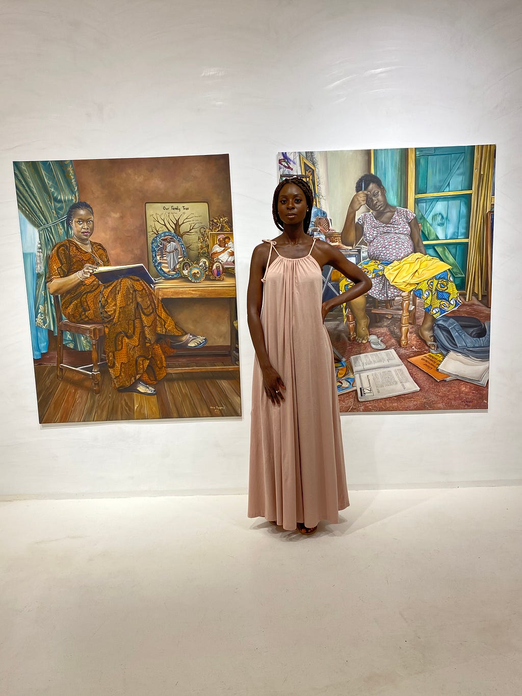 Woman standing in an art gallery in front of oil paint portraits. The portraits are of a woman sat in a chair reading a book, and the other is a woman sat with newspapers around her feet, an afro comb in one hand and batik fabrics drapped over her lap.