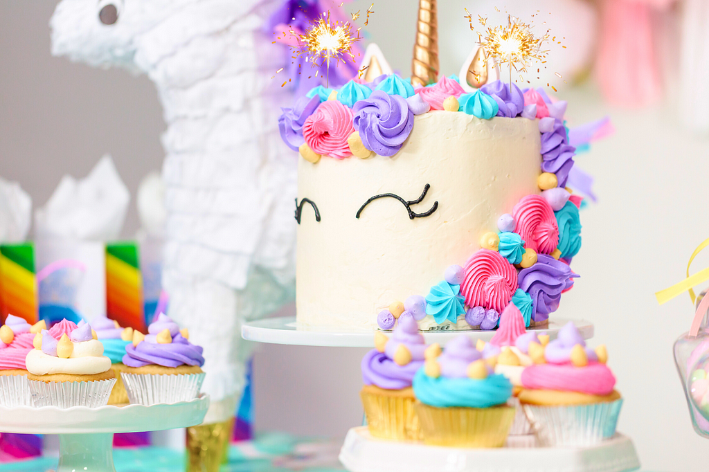A unicorn cake is surrounded by colorful unicorn cupcakes and rainbow decorations in the background. The cake has 2 sparklers on it.