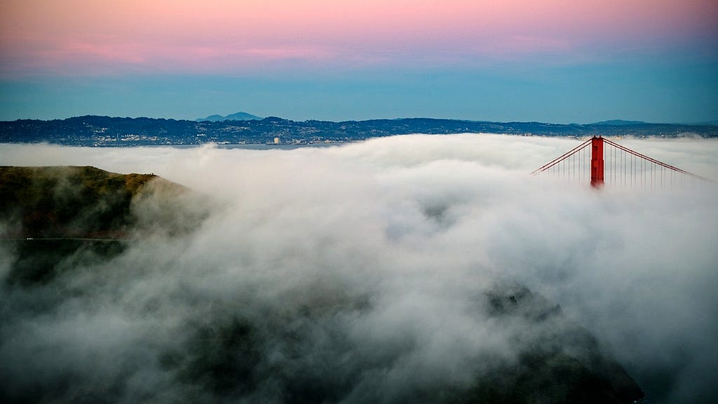 Foreground of fog in turmoil with tip of San Francisco Bridge showing above it. Background of rosy morning light on horizon.