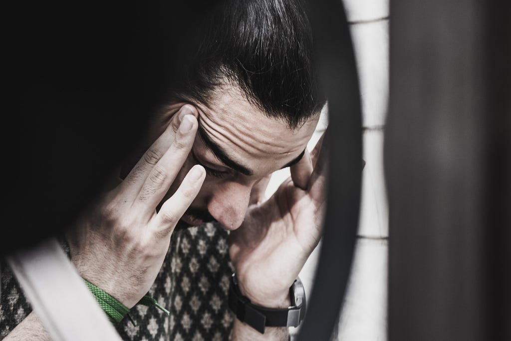 A man with brown hair and a mustache squeezing his temples looking like he’s in pain.