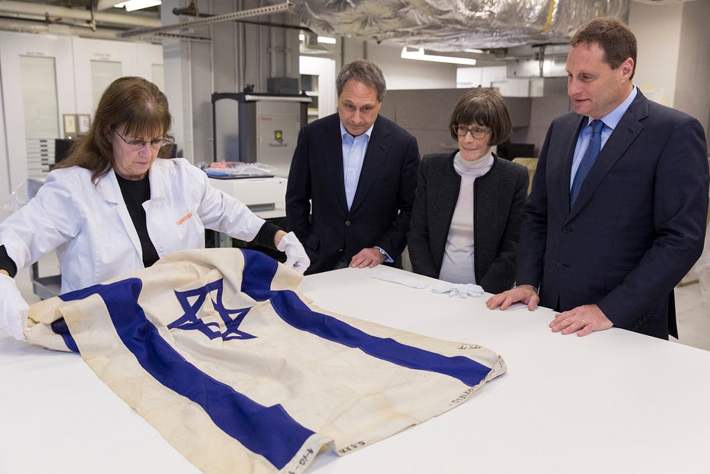 A woman wearing a white lab coat and white gloves unrolls a flag, while two men and one woman, all wearing black suit jackets, watch. The flag has a blue Star of David at the center and two horizontal blue cloth stripes near the top and bottom.