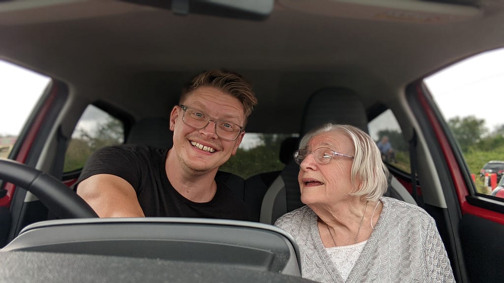 The author of the post and his mother sit in the front seats of a car. She looks at him lovingly. He looks directly at a camera on the cars dashboard.