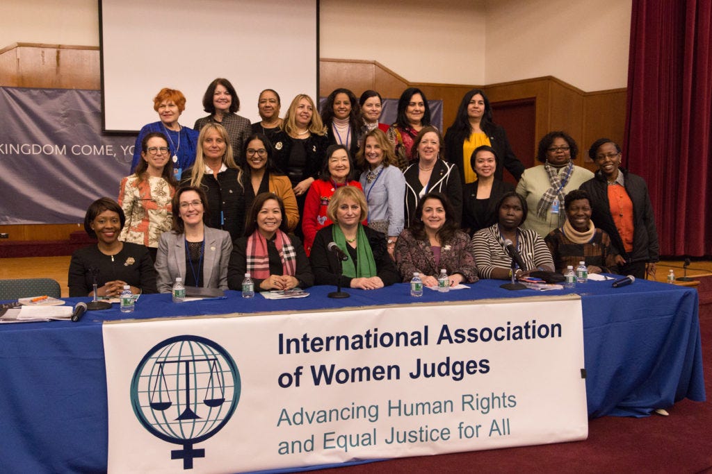 Adoley to the left, sitting on panel for International Association of Women Judges. With her hands collapsed on a blue table cloth covered table, Adoley is smiling and posing for picture with almost two dozen other women judges.