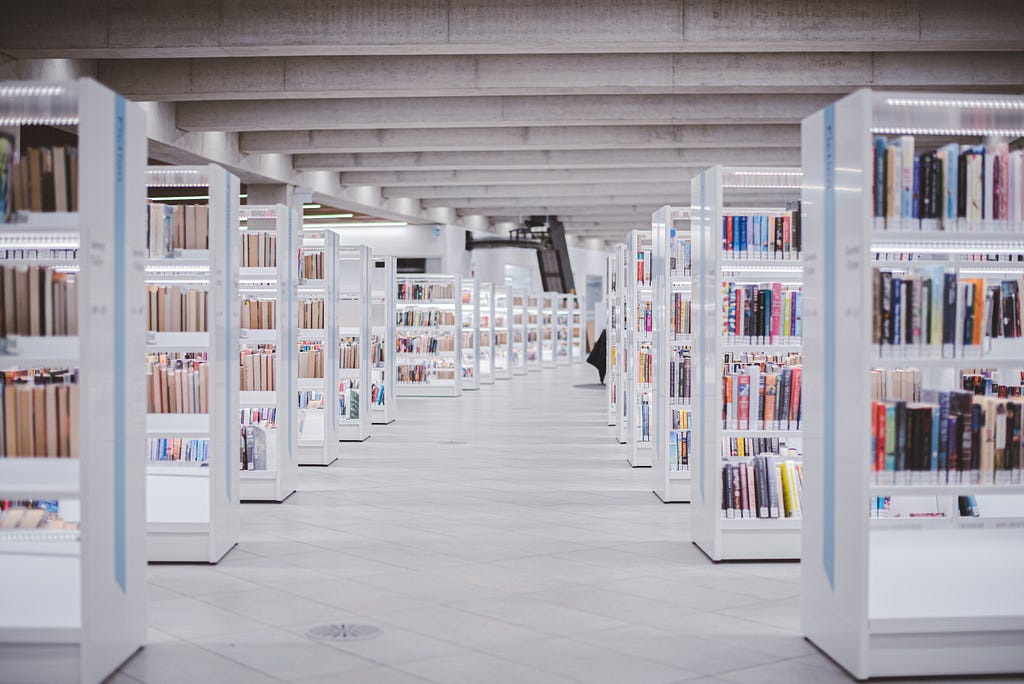 Rows of white, wooden bookshelves with books along either side of an aisle in a library.