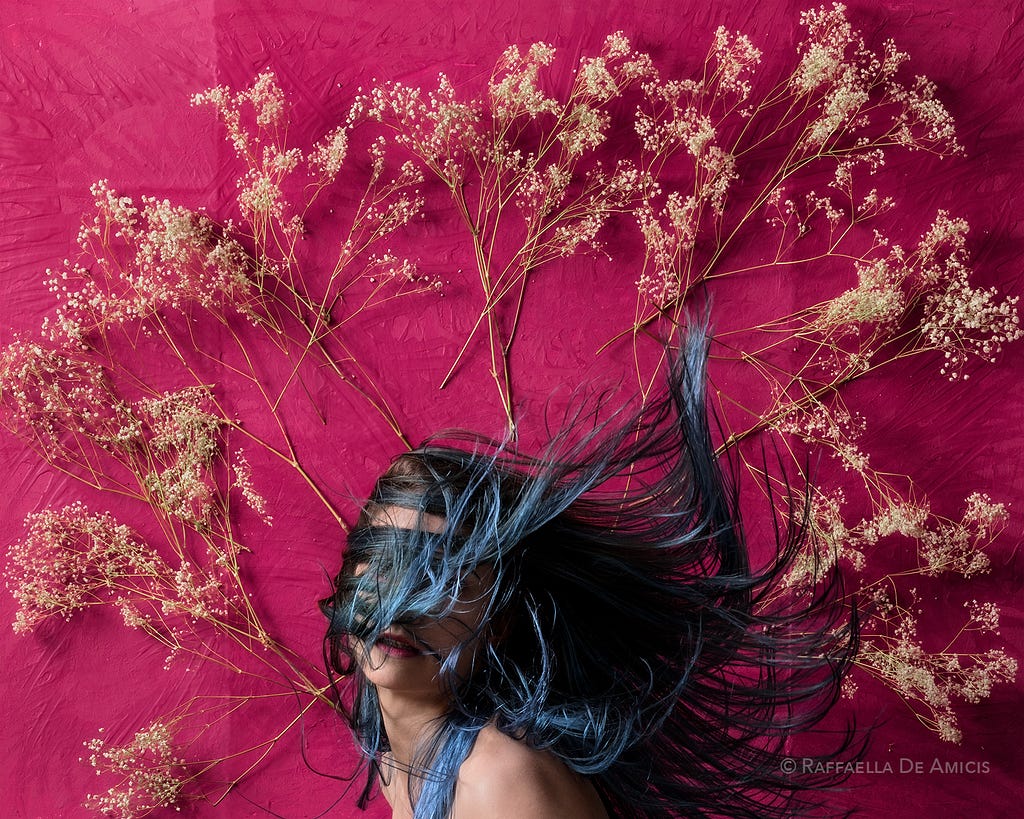 woman with blue hair flying in front of magenta background with dried flowers