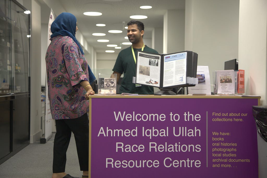 View of the welcome desk at the Ahmed Iqbal Ullah RACE Centre at Central Library in Manchester. A member of staff is welcoming a reader.