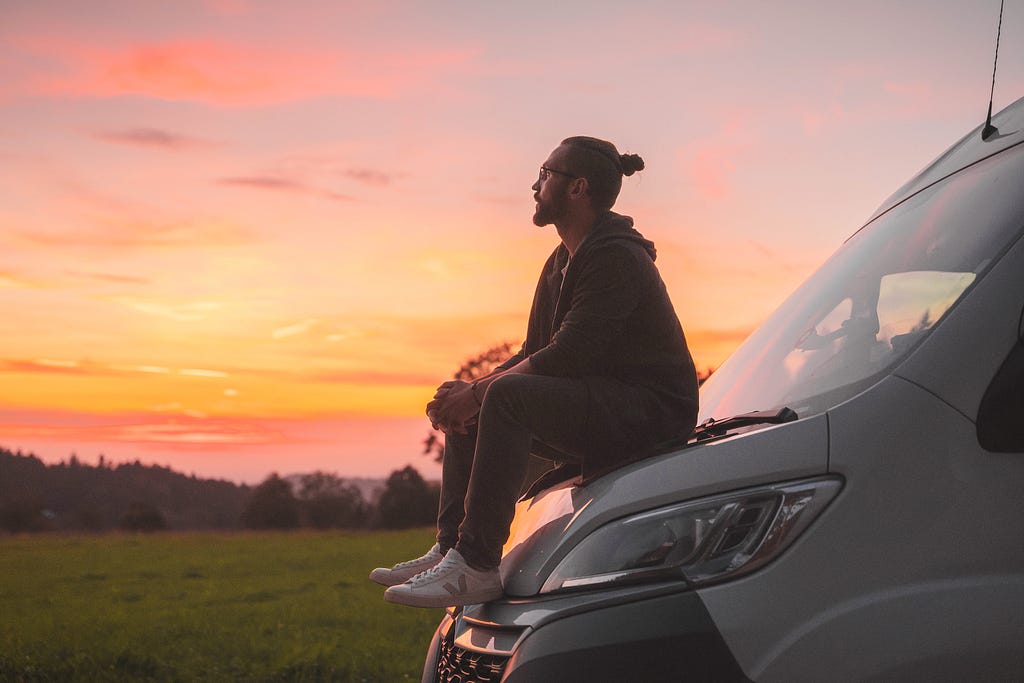 man sitting on front lid of a van during sunset