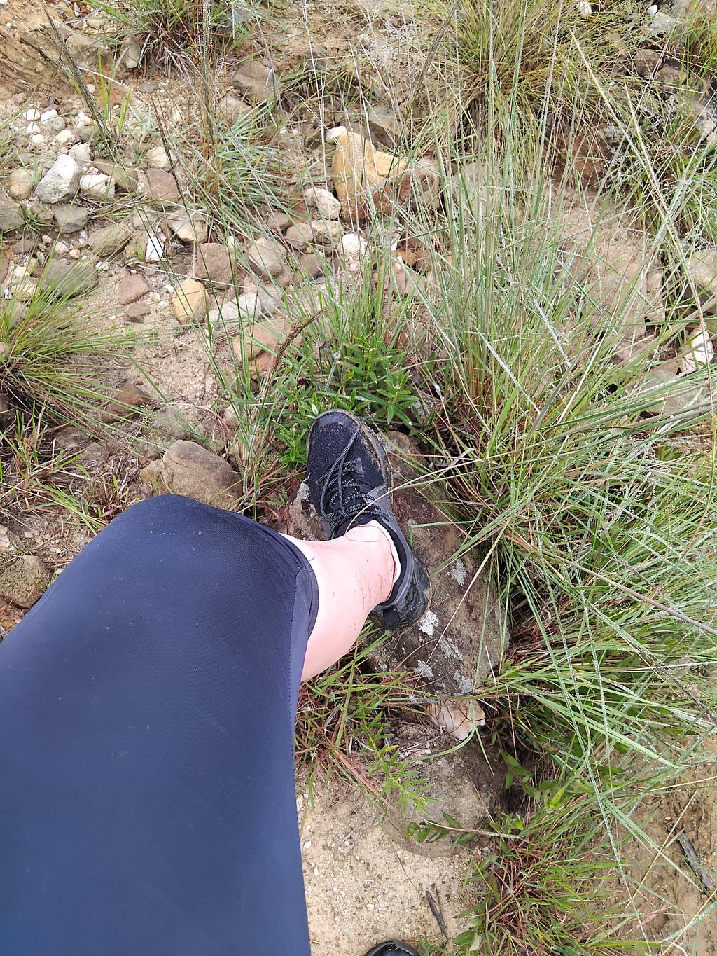 A Caucasian female leg wearing blue yoga pants and black shoes, resting on a rock, during a grueling hike in nature