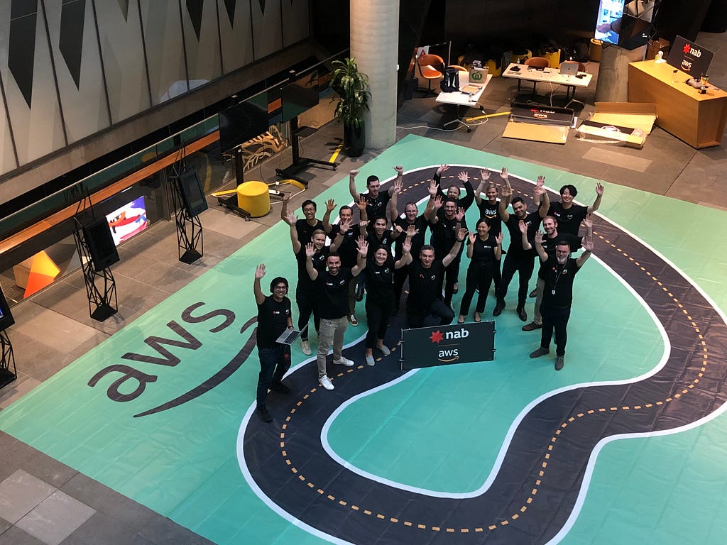 An aerial view of 18 people standing in the middle of the miniature-sized racing track and holding their hands in the air.