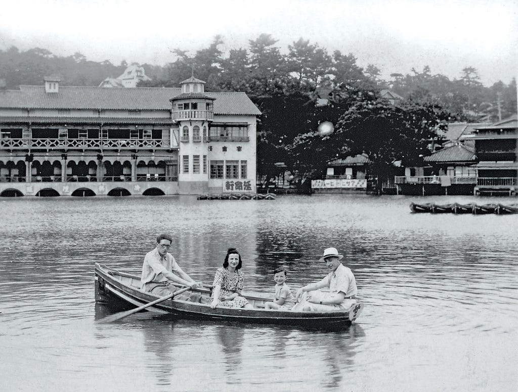 A man paddles a wooden row boat while a woman, a small boy, and a man in a white hat smile toward the camera. An ornate building with arches, balconies, and a turret is in the background. Behind it is lush foliage.