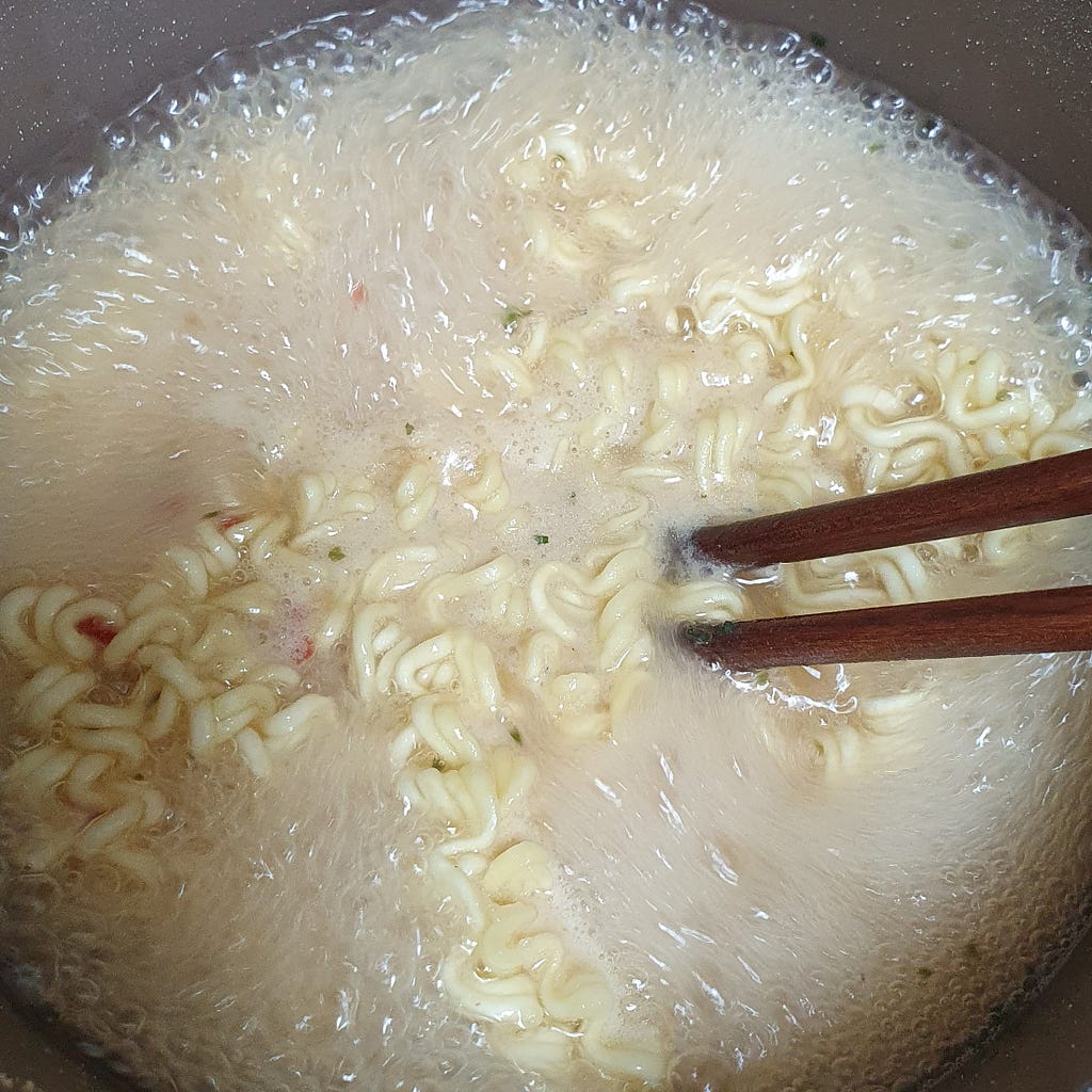 The ramen noodles and powdered soup packet from a package of Wicked Ramen (Yogoe Ramyeon) Vongole Flavor boiling together in the pot.