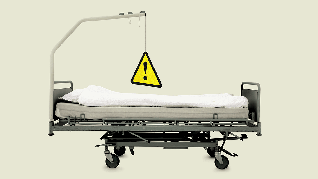 Side-view of a hospital bed with a metal bar above it; hanging from it is a triangular yellow caution sign.