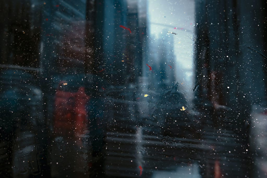 Abstract image of grey, dark grey, and red wash reminiscent of a blurred and rainy cityscape viewed behind dirty glass