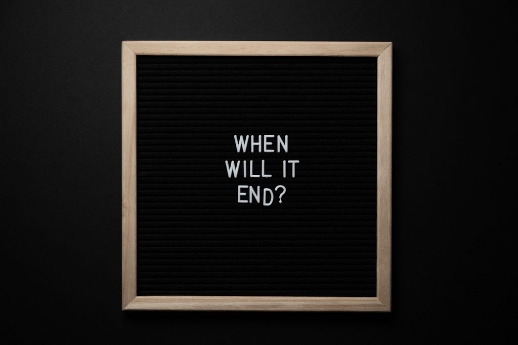 A wooden photo frame on black background. Inside, white letters spell the words “when will it end?”, underneath each other. The end tapers off as if falling from the frame.