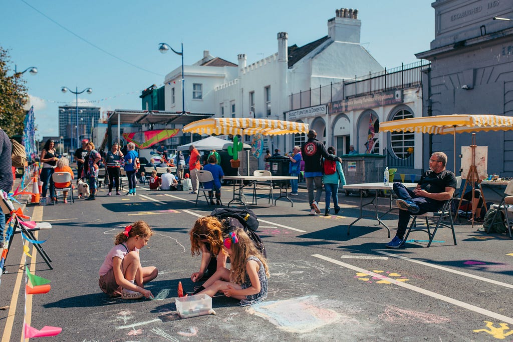 A photo of a lively and colourful street party in Plymouth. Children sit in a road drawing in front of marquees and tables.