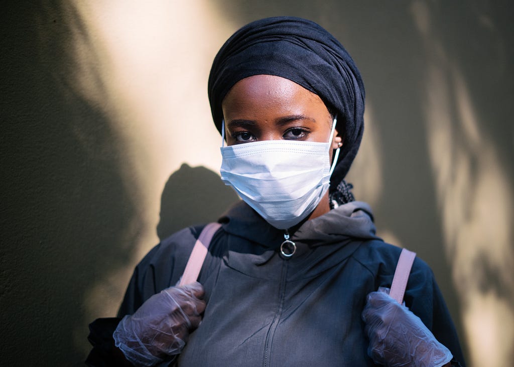 Black femme presenting person standing against the wall in shadows. She is wearing a mask and gloves with a blue jacket and backpack.