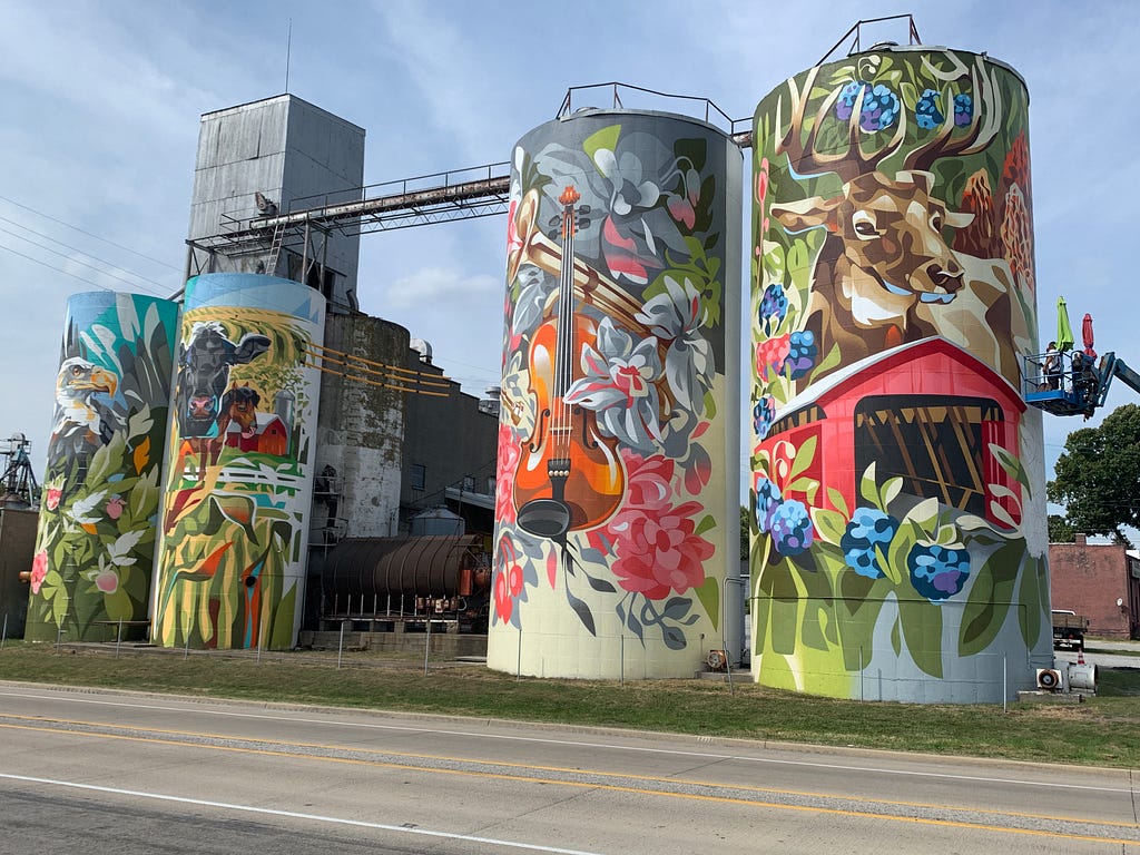Silos painted with colorful murals representing a region in Indiana with cows, barns, and musical instruments.