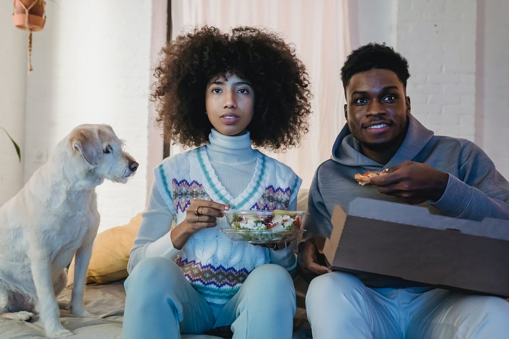 A focused young black couple having lunch and watching tv on the couch near the curious dog.