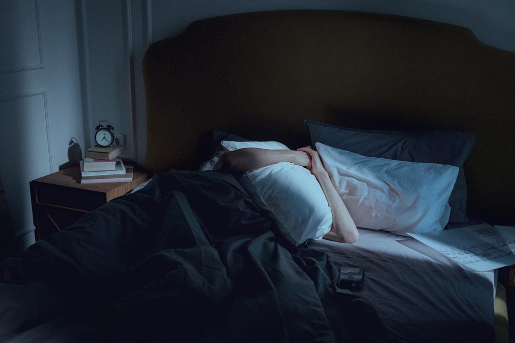 Man depressed in bed in dark room with pillow over face courtesy Ron Lach, Pexels (9615248)
