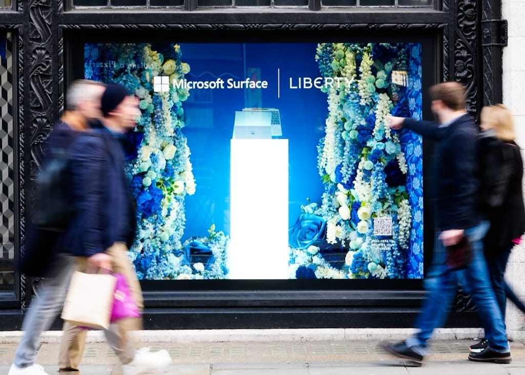 Image of people passing a blue storefront featuring a Microsoft Surface device on a podium against a blue backdrop with flowers
