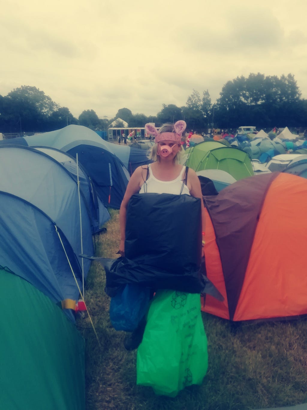 A white woman in her 30s in a field of tents wearing a pig snout and ears and a box covered in bin bags.