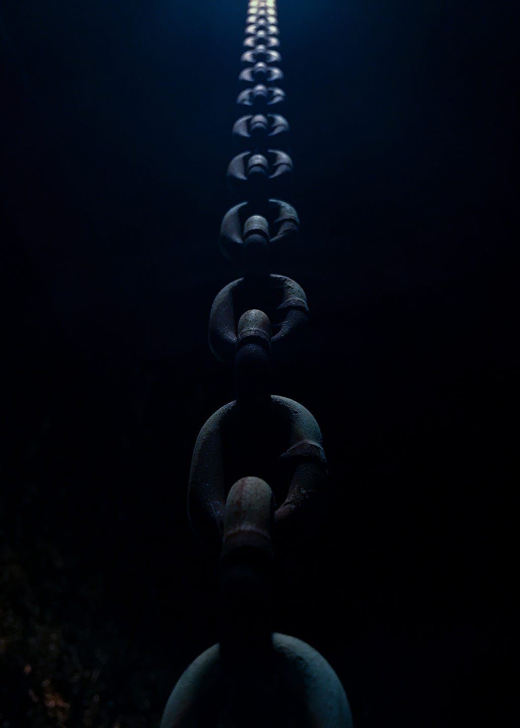 A dark space with a thick chain, adult hand size around big, taught leading down to a dry open, all dark surrounding, level below with an off of view light illumination of light above the chain.