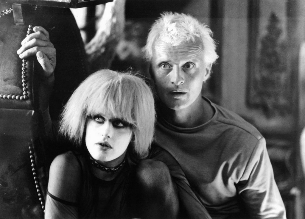 A female replicant, Pris, in Pierrot makeup and a male replicant, Roy, crouch next to each other.