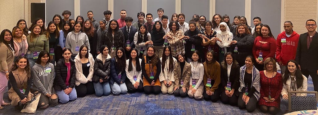 A group of youth lined up across a room to take a group photo. The photo depicts New American Youth Leadership Council (NAYLC) participants at the Youth Leadership Summit at the National Immigrant Inclusion Conference in National Harbor, Maryland in December 2022.