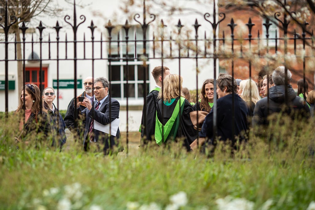 Graduates in their robes with their guests stand the other side of a metal fence.