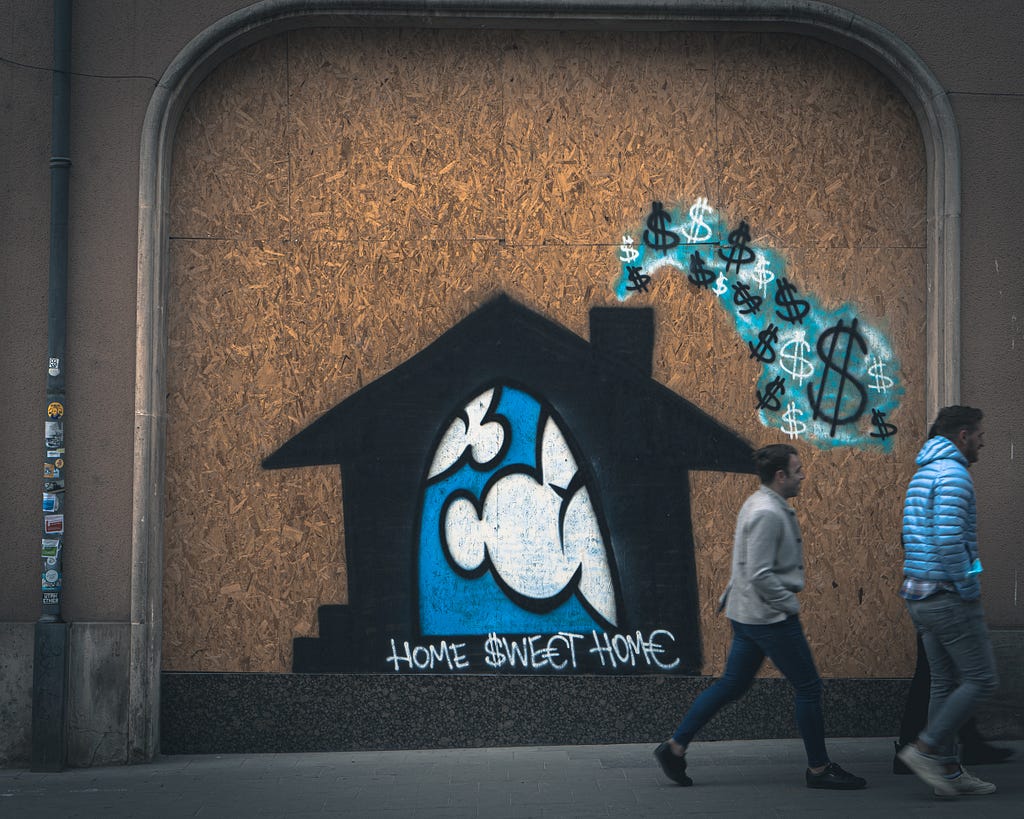 Graffiti art on a wall depicting a black cartoonish house with a chimney emitting dollar signs as smoke. Two men walk by the out of the frame to the right.