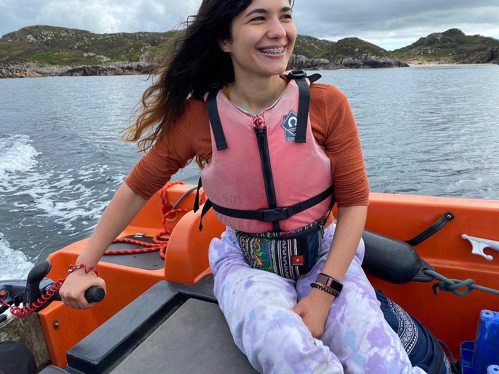 Irina smiling broadly and wearing a pink life jacket while steering a small boat across the water, with a background of rocky shoreline