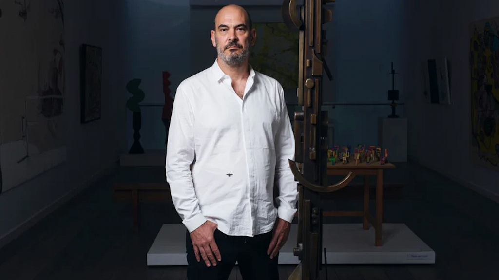 Andrew Klippel stands tall in a crisp white shirt, looking into the camera with hands poised at hips, surrounded by artifacts and art pieces in what might be a museum or gallery space.