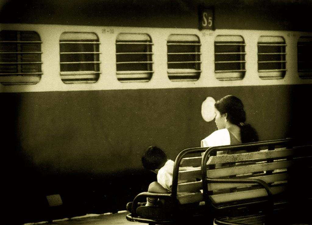 Monochrome image of a train at a station, with a mother and son on a platform bench.