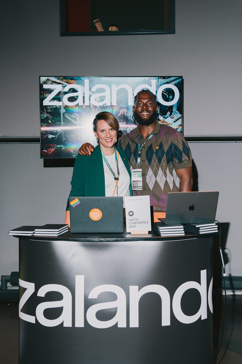 Two smiling people wearing conference lanyards and standing behind a Zalando-branded booth, with a large screen behind them and laptops and notebooks on top of the booth in front of them.