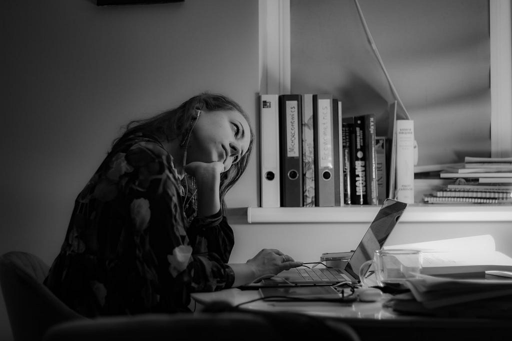 Candid photo of Tetiana Herasymova sitting at a table and working on a laptop with books and binders in the background. She has a forlorn look on her face and she’s resting her head in her hand.