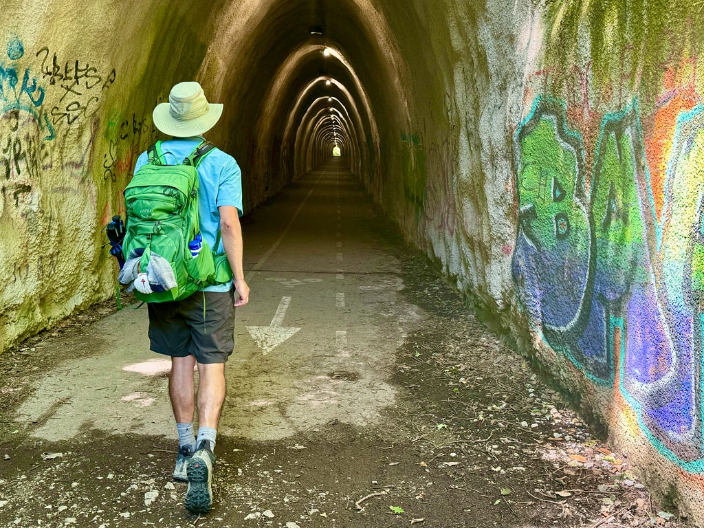 The author, with a green backpack on, entering a long (former) train tunnel that has graffiti on the walls.