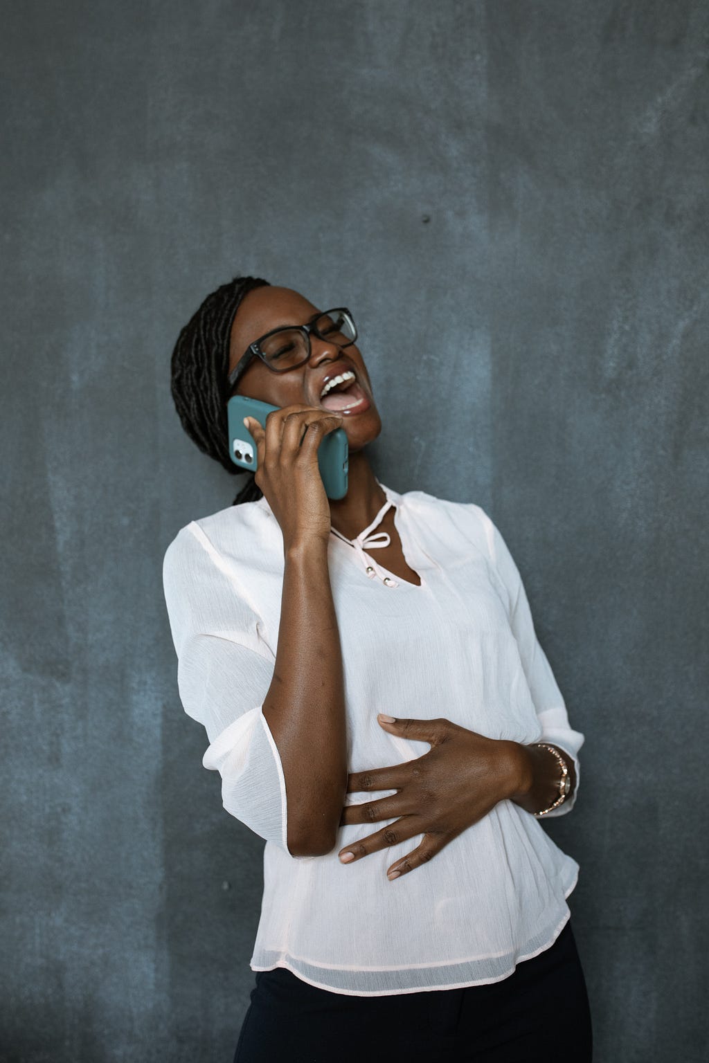 A woman in a white blouse is holding her phone laughing. The other hand is clasping her stomach.