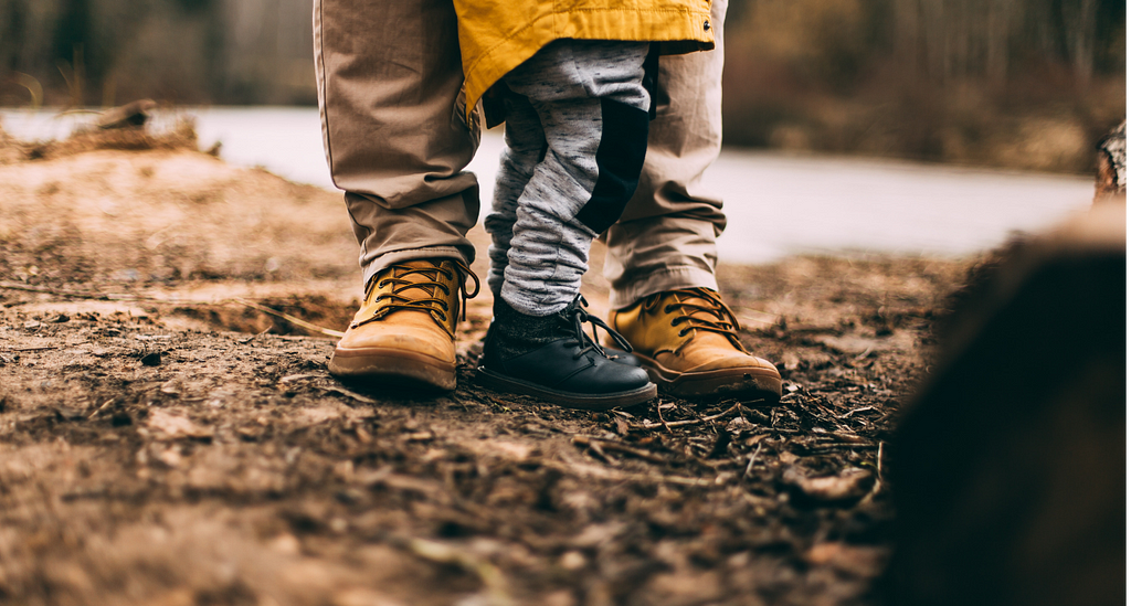 feet of a father next to feet of a small child standing on the ground both wearing warm-ish boots