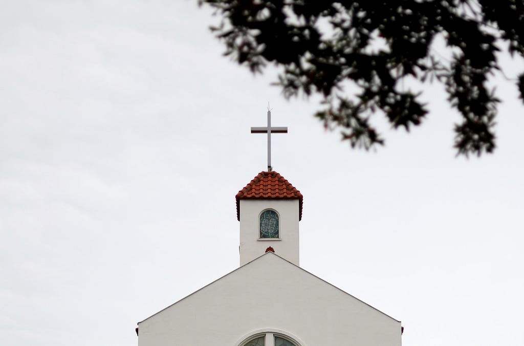 In the foreground at the top right are the unfocused branches of a tree. In the center of the image is the roof and steeple of a classic white church. It has an adobe tile roof and the steeple is ornamented with a simple cross at the top.