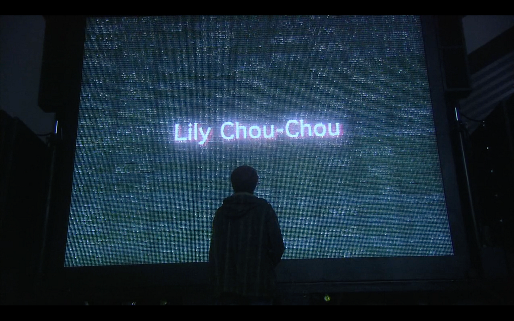 A teen boy’s black silhouette stands in front of a giant display screen, typically seen outside of concert venues, that has a dark blue-green glow. On the screen white text that spells out “Lily Chou-Chou” shines in the middle