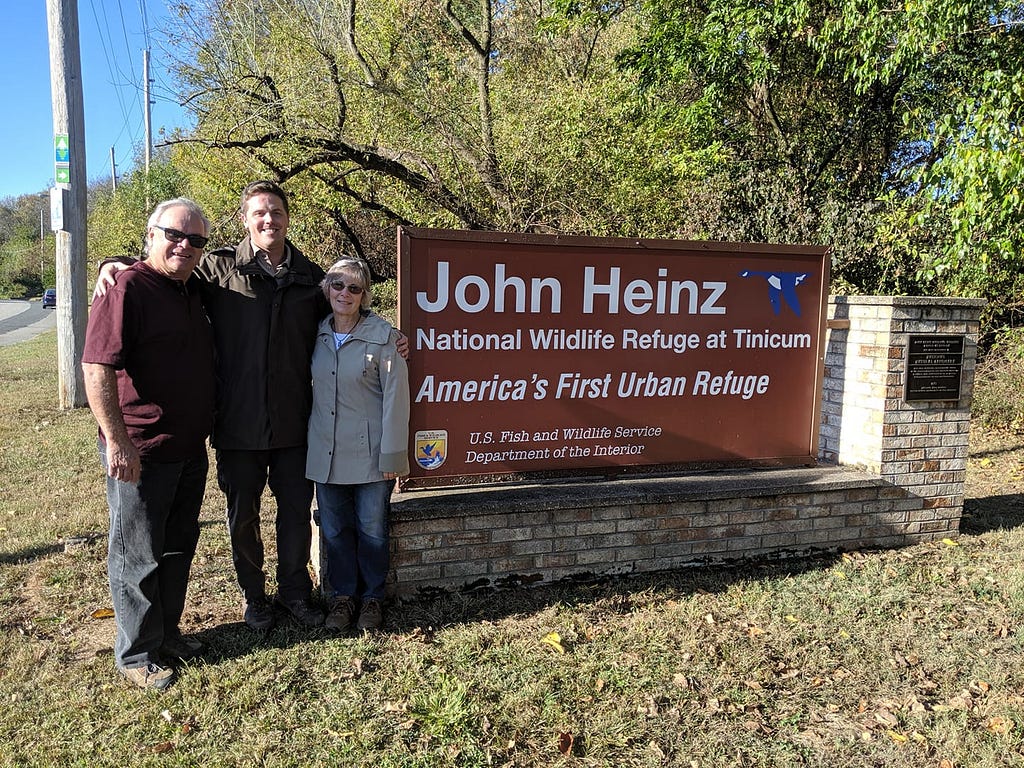 Kevin, David, and Cheryll stand together to the left of the John Heinz NWR welcome sign on a bright day.