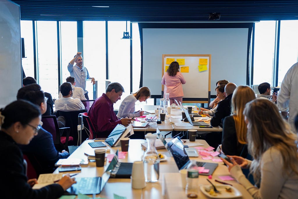 People sitting around a conference table working, as a person adds a sticky note to a white board at the end of the room. One person is standing speaking.